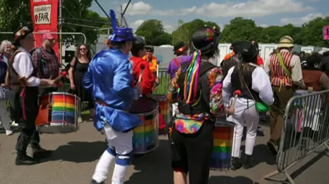 Several people are wearing colourful outfits and headpieces. They are all carrying and playing a drum while walking through an entrance.