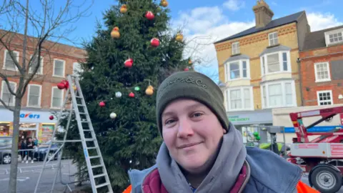 John Devine/BBC Annalise Bliss is standing in front of a partially decorated Christmas tree in a town. She is wearing a green woolly hat and a hoodie and jacket
