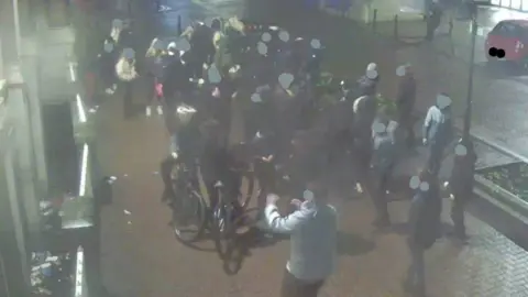 Grainy CCTV footage of hoard of young people in city centre. One has a bike. All their faces are blobbed out by grey dots. 