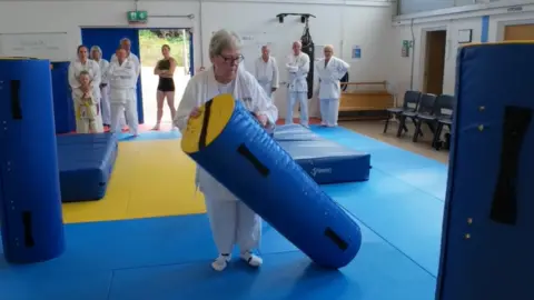 Harlow Judo Club A woman in white martial arts gear moving a blue, padded cylinder while her peers watch
