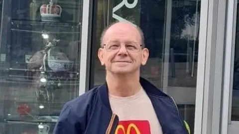 A man wearing glasses and a dark blue jacket is looking at the camera. He is standing outside a glass-fronted building with ornamental pottery works on shelves in the window.
