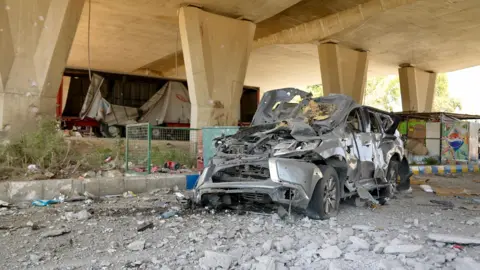 Goktay Koraltan / BBC A destroyed car in the Bekaa Valley, Lebanon - the car sits underneath a concrete overpass. It is surrounded by rubble and completely destroyed. 