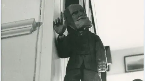 George Bernard Shaw Black and white photograph of a puppet dressed in a suit with one hand raised.