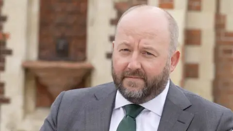 Nick Timothy is wearing a grey suit jacket on top of a white shirt and green tie.