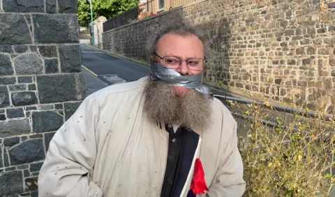 Lance Vaudin - A large man with a big bushy agrey and brown beard. Around his face across his mouth is silver gaffa tape. He's wearing rectangular rimmed glasses. he's also wearing a brown jacket.