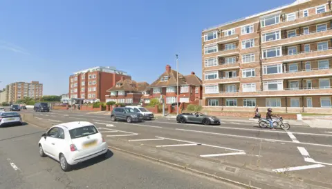 Google Two orange brick semi-detached houses sit by the side of a bus road between two large blocks of flats.