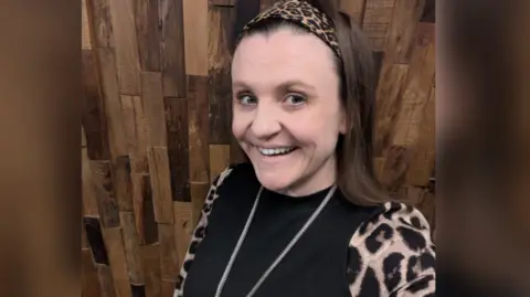 Tonia Dunn pictured in a selfie. She is smiling and stood against a wooden panel wall where she wears a leopard print sleeve shirt and silver long necklace.