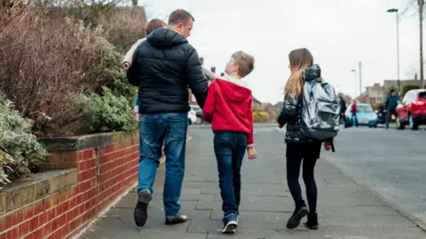 A father walks down a street holding a young child, with two other children walking alongside him.