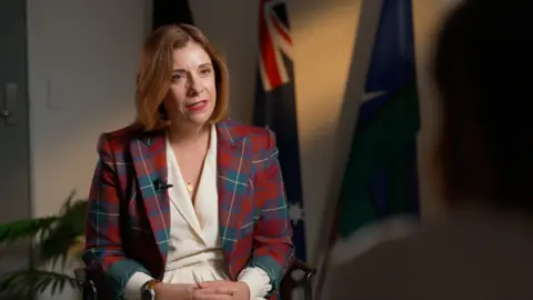 BBC A woman in a tartan jacket and white dress speaks. Several flags can be seen in the background