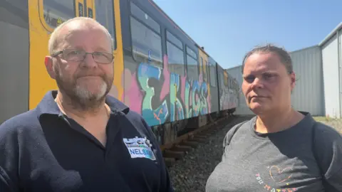 David Charles, who is wearing black thin-rimmed glasses and a navy blue polo shirt with the NELSAM logo of the museum, is standing next to Victoria Richardson, who is wearing a light grey shirt. They are both standing on ballast in front of the vandalised Metro car, which has been painted in pink, green and blue graffiti bubble writing spelling the word ERICS.