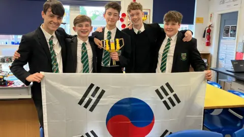 Five teenage boys, wearing black blazers, with one wearing a black jumper, over white shirts and green ties, smile as they hold a South Korea flag while standing in a science classroom. The boy in the middle is holding a golden Lego trophy.