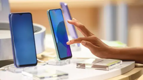 A young woman's hand touches the screen of a smartphone on display in a brightly-lit shop