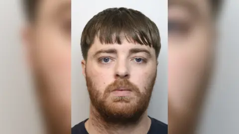 Wiltshire Police Callum Harvey-Mackenzie looking at the camera in a police mugshot. He has a long fringe and a full beard which is ginger.