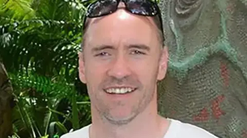 Paul Doyle, who has a shaved head, pictured smiling at the camera wearing a white t-shirt with sunglasses on the top of his head 