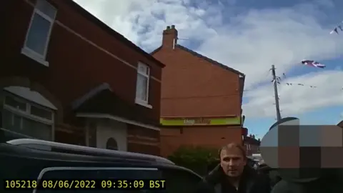 PSNI You can see two red brick buildings, one is a home, then other is a shop with Day Today signage on the side. 
A union flag can be seen flying in the background.
In the foreground, a man - Winston Irvine, it stood beside a car.
Beside him is a police officer - the PSNI has blurred his identity.
On the bottom left hand side of the picture there is yellow text which gives the date the footage was taken - the 8th of June 2022 - and the time - 9:35 am