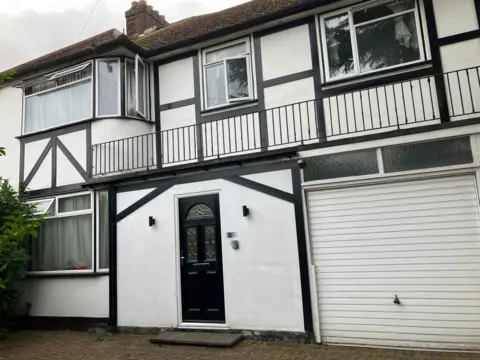 Brent Council A general view of the front of the house in Brent. In view we can see a black door, a white garage and some opened windows with white curtains.
