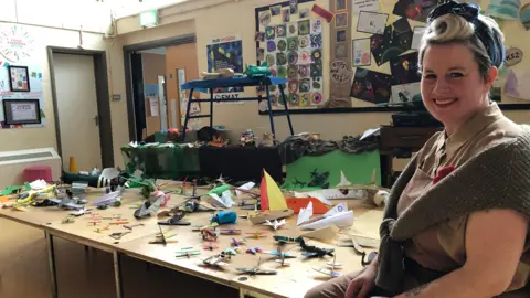 Clare Worden/BBC Head of Art at Nelson Academy Jill Sek dressed as a member of the Women's Land Army sitting in front of D-Day planes and boats made by the children. 