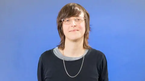 Getty Images A woman with shoulder length brown hair stands in front of a blue background. She is wearing a blue sweater, a long necklace and glasses. 