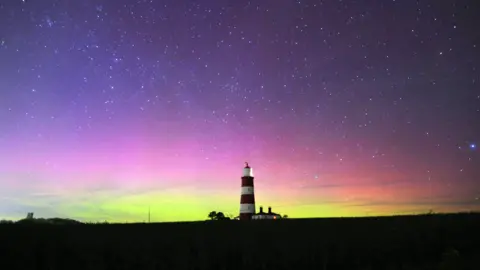 BBC Weather Watchers/KeepOnRunning A night-time landscape which has a striped lighthouse is in the middle. The Northern Lights have made the sky turn purple, pink, and yellow, and there are several stars.