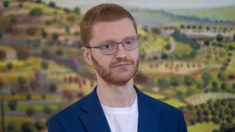 PA Media Ross Greer, who has short ginger hair and glasses, in a close-up shot. There is a picture of green fields behind him. 