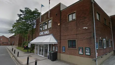 Stanley Civic Hall. It is a large two-storey red brick building with a set of white automatic doors. There is a canopy above the doors, and above this are large words on the side of the building that say Civic Hall. There are also two flag poles at the top of the building.