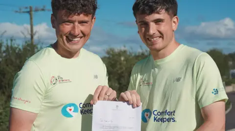 Laura-Mary Carter/FTD Brothers Two men in light green T-shirts are holding a letter. They are looking at the camera and dominate the image, with bushes in the background.