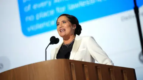 Getty Images Stephanie Grisham speaks at a podium wearing a black shirt and white jacket