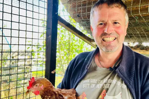 A man holds a hen in a coop on an allotment in Hessle. He is smiling and wearing a blue cardigan and a grey T-shirt.