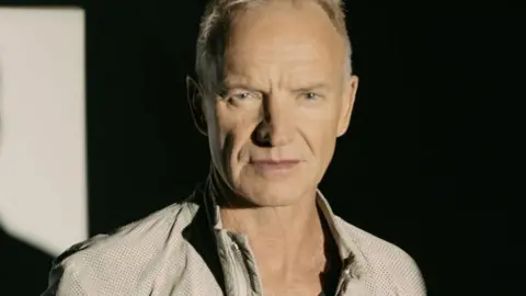 Sting in a casual beige jacket looking thoughtful. His is standing against a black background and to the left part of the wall is lit up. 