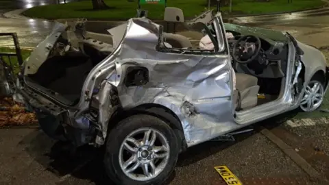 A badly damaged silver car, half on the pavement at a round-about in the dark.  The roof is missing.