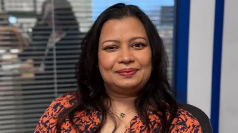 Anindita Sarkar is sitting in a chair. She has long brown hair and is wearing a blue and orange dress. Out of focus behind her is an office.