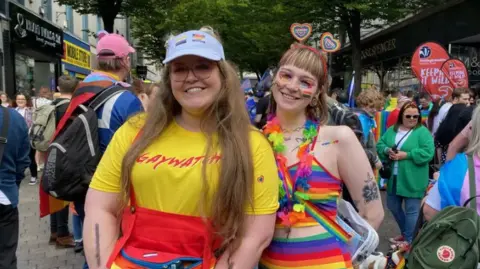 Two woman at a recent pride march in Nottingham. One woman is wearing a yellow t-shirt with GAYWATCH on the front. Another woman next to her is wearing a rainbow-coloured dress with rainbow makeup and a floral necklace. People are milling in the background.