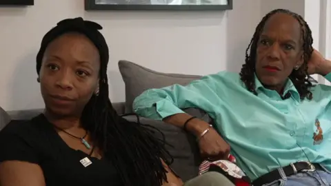 TJ Jacobs, wearing a black t-shirt and a necklace, is sitting on a grey sofa next to Vyleen Maximen, who is wearing a mint green shirt, black belt and blue jeans