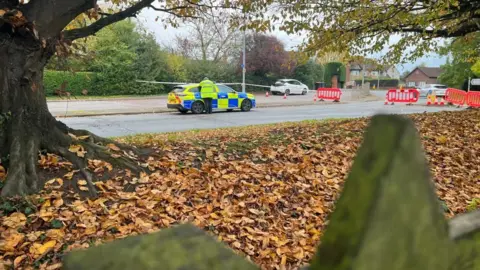 Emma Baugh/BBC A police car, with a policeman standing by the side, in a road that has been cordoned off. The cordon is in the distance, with leaves in front. The cordon shows red fencing and cones. 