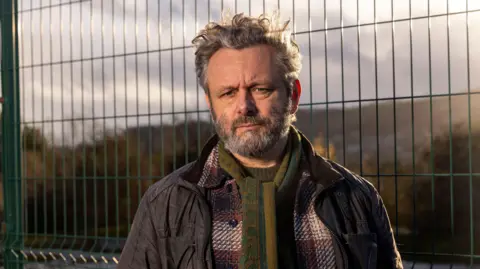BBC/Wall to Wall Media/Stephen Perry The image is a close outdoor portrait taken near a wire-mesh security fence. Michael Sheen stands facing the camera, dressed for cool weather in several layers, including a dark field jacket, a checked wool coat and a patterned green scarf. The fence fills much of the background, while distant hills and trees are softened by shallow depth of field. Golden sunlight from a low angle adds warmth to the scene and emphasises the textures of the scarf, coat and fence.