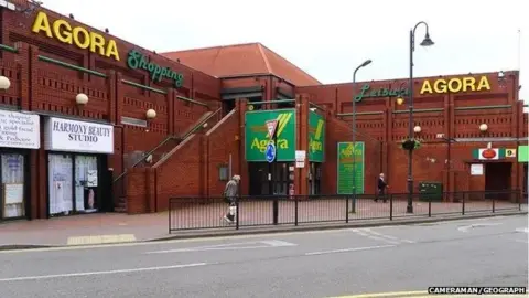 CAMERAMAN/GEOGRAPH Red-brick commercial building with Agora in yellow writing on the top edge. There are a few shop units remaining including a beauty parlour and post office. There is black railing separating the building from the road that runs alongside it.