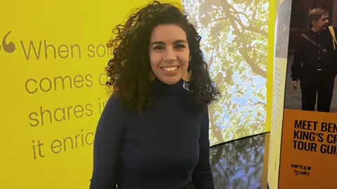 Gem O'Reilly A woman with brown curly hair and wearing a long-sleeved black top and yellow earrings stands in front of exhibition billboards. 