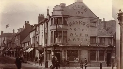 Black and white archive picture of a pub, with Tiger Inn painted on the eaves, under which further painting reads "Banks's noted ales and stouts". People mill around on foot on the streets outside.