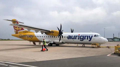 A propeller-powered plane on a runway, with Aurigny livery. A man in a high-vis jacket moves an orange cone.
