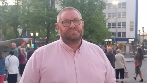 A man, with short dark hair and a tight beard, wearing a pink shirt with rolled up sleeves. There are people standing around behind him in the background.