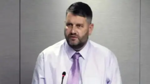 Alan Paton, a bearded man in a purple shirt and tie is sitting in front of a grey wall.