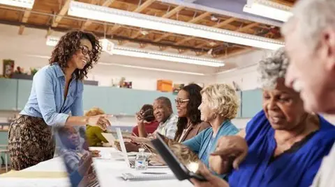 A group of elderly people sitting inside a community centre on laptops and talking to each other and a woman with dark hair who stands in front of them.