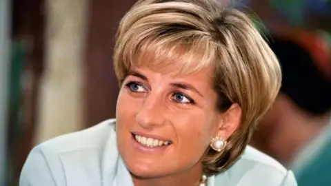 Princess Diana looks upwards at an angle smiling while a photographer takes her picture.