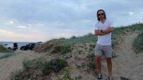 Councillor Tristan Learoyd standing on a sand dune at Marske beach with two cars parked in the distance, close to the shore. He has shoulder-length brown hair and is wearing dark sunglasses, a white t-shirt, beige shorts and black and white skater shoes. He has his arms crossed and is looking at the camera with a serious expression.