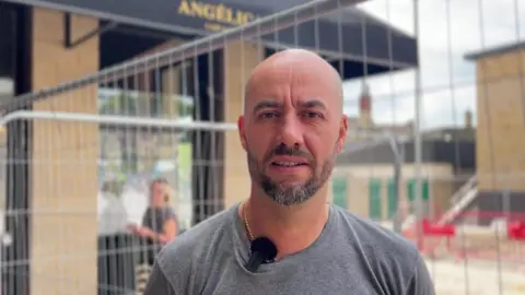 NATHAN TURVEY/BBC A picture of Blair Manovi who has a beard and is wearing a grey top, standing in front of his bistro with metal railings and roadworks behind him.