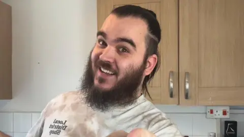 Craig Fitzpatrick Craig lleva una camiseta blanca y sonríe y señala a la cámara contra un armario de la cocina.  