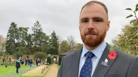 Peter Saunders looks at the camera, wearing a grey suit with blue tie and shirt, wearing a poppy and Royal British Legion badge. He has ginger hair and beard and is in a field of remembrance with people and wooden crosses.