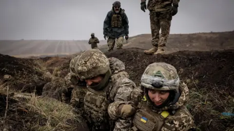 EPA Ukrainian soldiers, in camouflage military clothing, stand in a trench during a training exercise. Behind them, more personnel are standing in an otherwise empty, brown field.