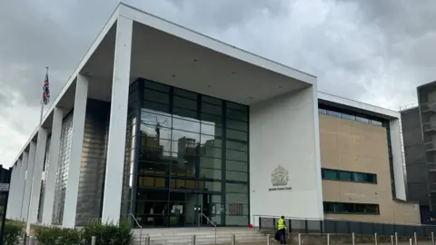 The front of the Ipswich Crown Court building. It is a cloudy day. A person in a high-vis yellow jacket appears to be tidying up outside the court entrance.