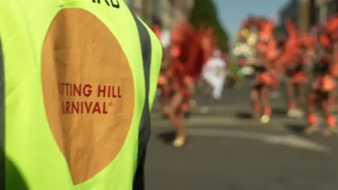 A steward wearing a Hi-vis yellow jacket with Notting Hill Carnival written on it stands in the foreground, with a scene of dancers in colourful red costumes in front of him